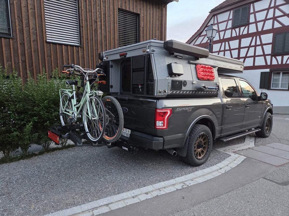 CraneRock TP03 auf Ford F-150 vor einem Schweizer Riegelhaus in der Abenddämmerung, mit E-Bikes am Heckträger